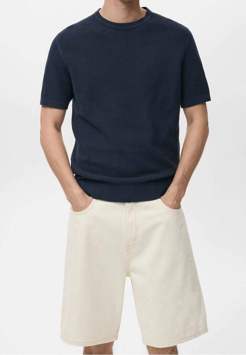 Homme portant un pull à manches courtes texturé bleu marine foncé et un short beige clair longueur genou, les mains dans les poches.