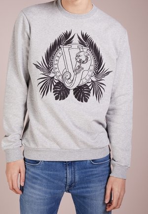 Persona che indossa una felpa grigia con foglie tropicali nere e un grande logo V decorato con disegno di leone, abbinata a jeans blu.