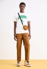Hvid T-shirt med grøn "JORDAN" tekst, brune bukser og beige sneakers med brune accenter. Lille brun rund taske med en rem.
