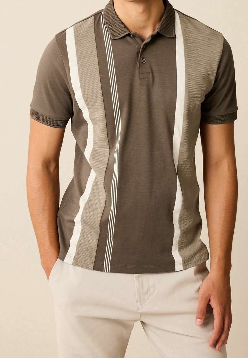 Bruin poloshirt met witte gestreepte accenten, voorzien van een gestructureerd ontwerp aan de zijkanten en een klassieke kraag. Gedragen met lichtgekleurde broek.