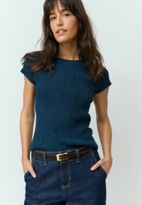 Strickoberteil in Teal mit gerippter Textur, kurzen Ärmeln und rundem Ausschnitt, kombiniert mit dunklen Denim-Hosen und einem braunen Gürtel.