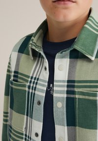 Zeleno-bež karirasta srajca s gumbastim ovratnikom, ki vsebuje temne gumbe, žepom na levi strani prsnega dela, preplastena z temno modro majico.