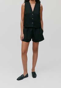 Czarna lniana kamizelka z dekoltem w serek i pięcioma guzikami z przodu, zestawiona z pasującymi czarnymi szortami. Nosi się z teksturowanymi czarnymi butami typu slip-on.