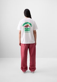 T-shirt bianca in cotone con una grafica colorata sul retro, caratterizzata da testo verde e rosso, indossata con pantaloni rossi larghi.