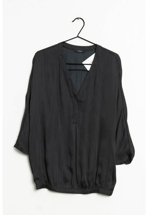 Sort bluse med lange ærmer, V-hals, løs pasform og elastisk kant, hængende på en sort bøjle mod en hvid baggrund.