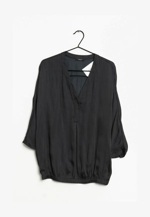 Sort bluse med lange ærmer, V-hals, løs pasform og elastisk kant, hængende på en sort bøjle mod en hvid baggrund.