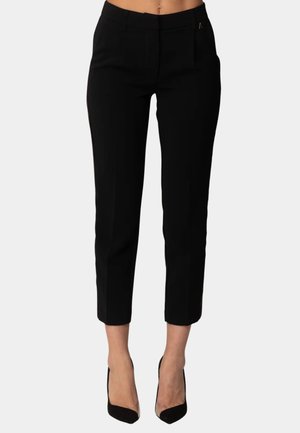 Donna che indossa pantaloni neri slim-fit cropped abbinati a scarpe nere con tacco alto, mostrata su uno sfondo chiaro e uniforme.