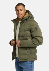 Green puffer jacket with a soft, beige fleece lining, zip closure, hood, and zipper pockets. Features stitched panel design and logo patch.