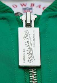 Grüner Stoff mit einem silbernen Metallreißverschluss-Zuggriff, der den Text "Mitchell & Ness" und "Nostalgia Co. Philadelphia, PA." trägt.