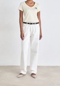 Camiseta de algodón amarilla pálida con escote en V y logo, combinada con pantalones de mezclilla blancos de corte holgado y bailarinas puntiagudas gris claro. Cinturón de cuero negro.