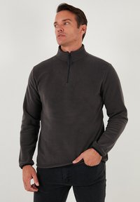 Mørkegrå fleecepullover med halv-zip krave, lange ærmer og en blød tekstur. Subtil syning detalje langs kanten og manchetterne.