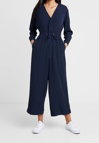 Mujer vestida con un mono de manga larga azul marino con cintura ajustable y zapatillas blancas, de pie con las manos en los bolsillos.