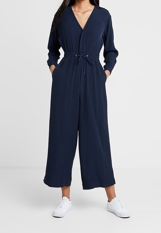 Mujer vestida con un mono de manga larga azul marino con cintura ajustable y zapatillas blancas, de pie con las manos en los bolsillos.