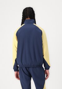 Veste de survêtement marine et jaune avec un col montant, des poignets élastiques et une coupe décontractée. Présente un bloc de couleur contrastant sur les manches.