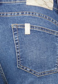 Jeans de mezclilla azul con un bolsillo trasero que presenta una etiqueta blanca con la inscripción "L.J." y costuras amarillas contrastantes. Textura suave y diseño clásico.