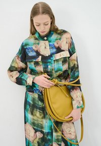Bloemmotief blouse in groen, blauw en roze, met knoopsluiting en twee voorzakken. Houdt een ronde gele leren handtas met een handvat vast.