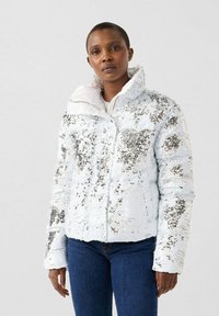 Woman wearing a white sequined puffer jacket with high collar and blue jeans, standing against a plain light background.