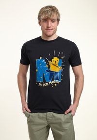 Svart bomulls t-shirt med en färgglad grafisk design med en gul björn och en blå trollkarlshatt. Texten säger "M ÄR FÖR MAGI!" med stjärnor.