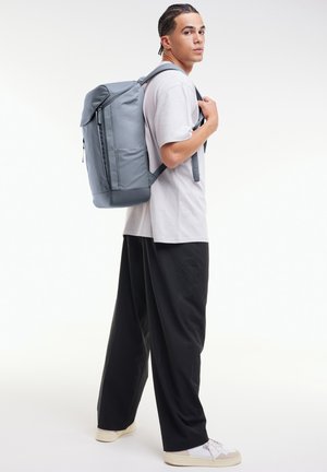 Mochila gris con un diseño aerodinámico, superficie texturizada, bolsillo frontal con cremallera y correas ajustables, llevada por una persona vestida de forma casual.