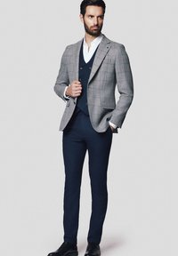 Szary w kratkę żakiet na granatowej kamizelce, zestawiony z granatowymi spodniami. Mężczyzna stoi pewnie, mając na sobie czarne buty. Gładki materiał i dobrze dopasowany krój.