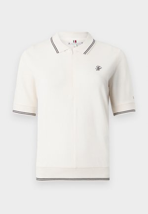 Polo color marfil en un material de punto suave. Cuenta con un cuello corto con rayas azules marinas, cierre de cremallera y un logo bordado en el pecho.