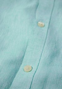 Jasnoturkusowa koszula zapinana na guziki, wykonana z teksturowanej tkaniny, z jednym okrągłym, jasnym guzikiem wzdłuż listwy, o gładkim wykończeniu.