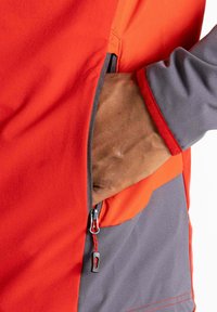 Rote und graue Jacke mit einer Reißverschlusstasche, strukturiertem Stoff und kontrastierendem Akzent um die Taschenöffnung. Die Hand ist in der Tasche.