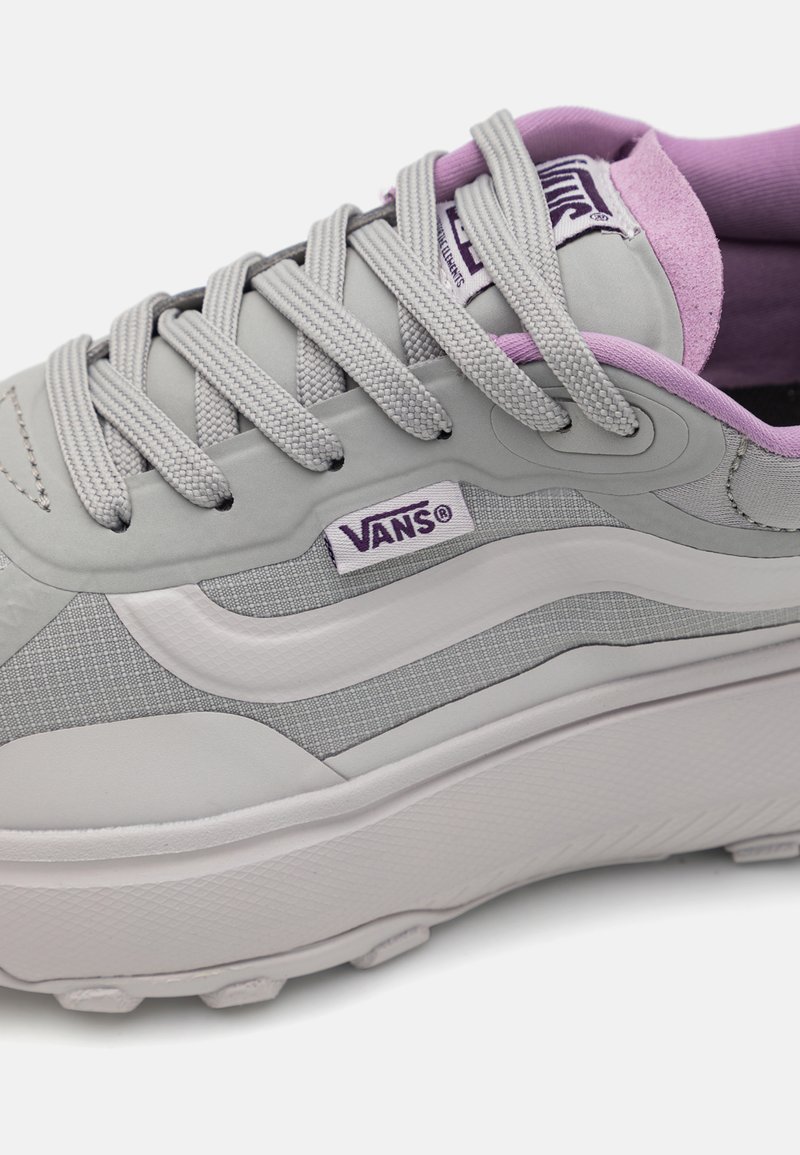 Zapatilla sintética gris con acentos lilas claros. Presenta una superficie texturizada, un diseño de onda blanca prominente y una suela de goma con patrón de tracción.