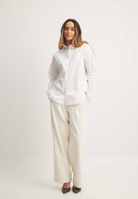 Witte overhemd met lange mouwen, gecombineerd met textuurloze, losse crème broek en grijze hakken. Eenvoudig, schoon ontwerp.
