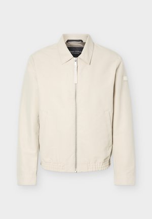 Veste beige à fermeture éclair avec col, manches longues, deux poches latérales, taille élastique et un petit patch sur la manche gauche.