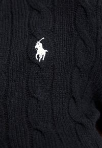 Svart kabelstrikket genser med en hvit brodert polo-logo. Stoffet har et teksturert mønster med hevet kabelstrikk.