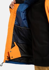 Osoba w czerni pokazuje wnętrze otwartej pomarańczowej kurtki z niebieską podszewką i czarną siatkową kieszenią.