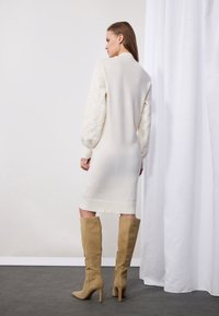 Robe en tricot ivoire avec des manches en point de câble texturé, présentant une silhouette longue et un ourlet côtelé. Associée à des bottes en suède beige montantes.