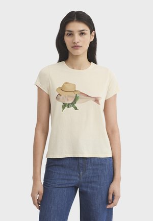 Femme portant un t-shirt crème avec un motif d'un poisson portant un chapeau de paille et une écharpe verte, associé à un jean bleu.