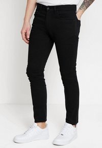 Vaqueros negros ajustados de mezclilla, con un diseño clásico de cinco bolsillos y cierre de botón. Combinados con zapatillas blancas para un contraste.