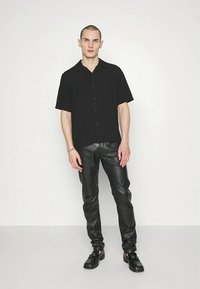 Black button-up short-sleeve shirt, paired with tight-fitting black leather pants. Model stands against a plain white background.
