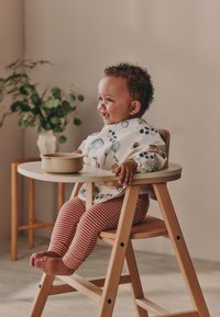 Chaise haute avec une structure en bois clair, équipée d'une surface de table ronde. L'enfant porte un bavoir blanc avec des motifs colorés et un legging rayé rouge.