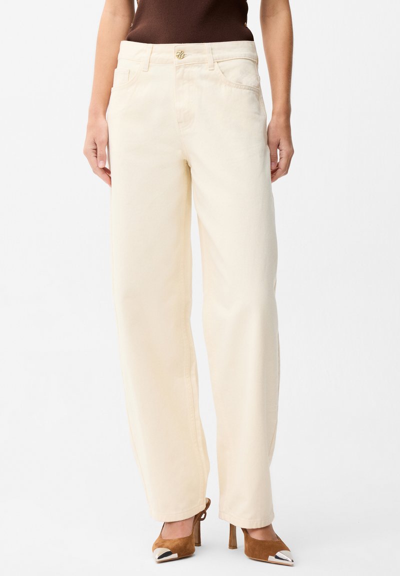 Femme portant un pantalon droit écru, un haut marron sans manches et des escarpins marron en daim avec des bouts métalliques.