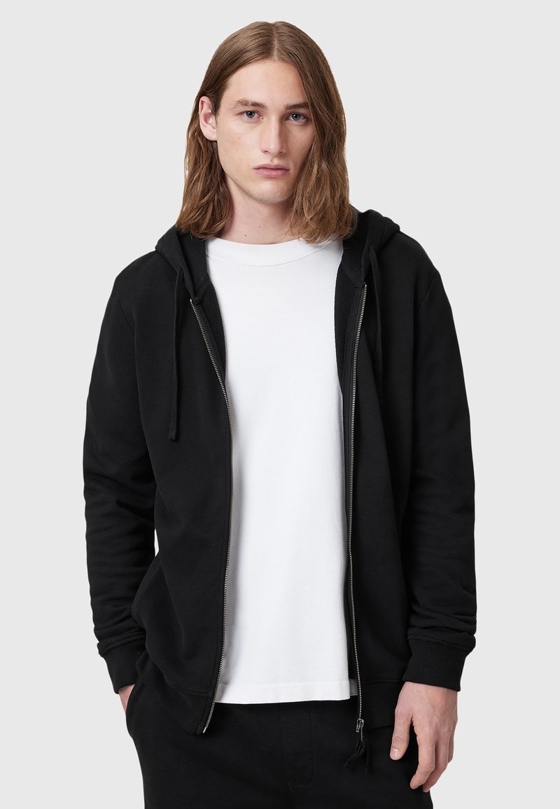 Hoodie noir zippé avec capuche à cordon, présentant une texture lisse et des poches latérales, superposé sur un t-shirt blanc uni.