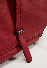 Červená kožená taška s hladkou texturou, vybavená plochým popruhem a stříbrným kovovým akcentem. Minimalistické šití podél okrajů.