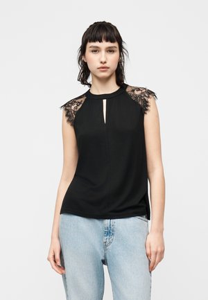 Ung kvinde i sort ærmeløs top med blonde detaljer på skuldrene og lyseblå jeans, stående mod en ensfarvet hvid baggrund.