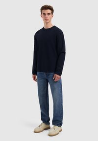 Marineblauwe lange mouwen shirt van katoen met een ronde hals, gecombineerd met lichtblauwe denimjeans en beige schoeisel, tegen een neutrale achtergrond.