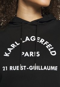 Černá mikina s kapucí a bílým textem. Mezi její vlastnosti patří šňůrka, měkká textura a výrazné logo: "KARL LAGERFELD PARIS 21 RUE ST-GUILLAUME."