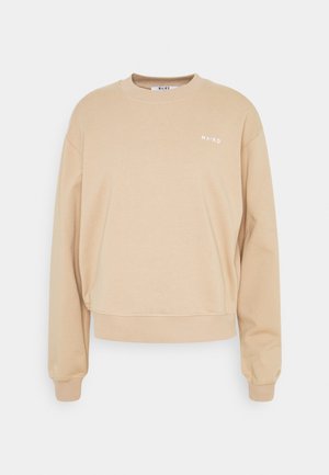 Sudadera beige con cuello redondo, hombros caídos y puños acanalados. Presenta un pequeño logotipo blanco de "NA-KD" en el pecho. Textura de tela suave.