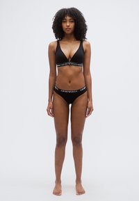 Σετ μαύρου σουτιέν και μπικίνι με σχεδιασμό σε σχήμα V. Διαθέτει λευκές λωρίδες με το λογότυπο της μάρκας που αντιπαραβάλλονται γύρω από τη μέση και κάτω από το στήθος. Ομαλή υφή.