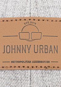 Harmaa kuviollinen kangas, jossa on ruskea nahkamerkki, jossa on kohokuvioitu teksti. Merkissä lukee "JOHNNY URBAN" ja "METROPOLITAN ACCESSORIES."