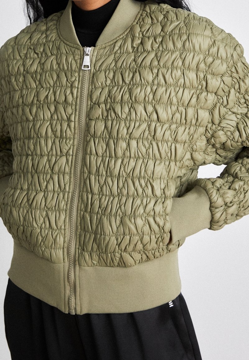 Personne portant un blouson bomber vert olive texturé avec fermeture éclair, poignets et taille côtelés, mains dans les poches ; pantalon noir visible.