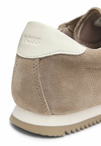 Beżowe zamszowe sneakersy z białym skórzanym akcentem przy pięcie, wytłoczonym logo "BOSS" oraz szarym gumowym spodem z ząbkowaną krawędzią.