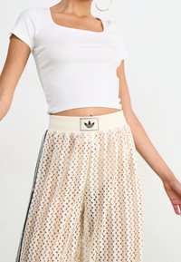 Valkoinen sovitettu lyhyt toppi, joka on yhdistetty vaaleanbeigeihin, teksturoituihin leveälahkeisiin housuihin, joissa on ziksak-kuvio ja Adidas-logo vyötäröllä.