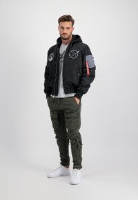 Giubbotto bomber nero con cappuccio, toppe ricamate, chiusure con cerniera, abbinato a pantaloni cargo oliva e sneakers bianche. Sfondo neutro.
